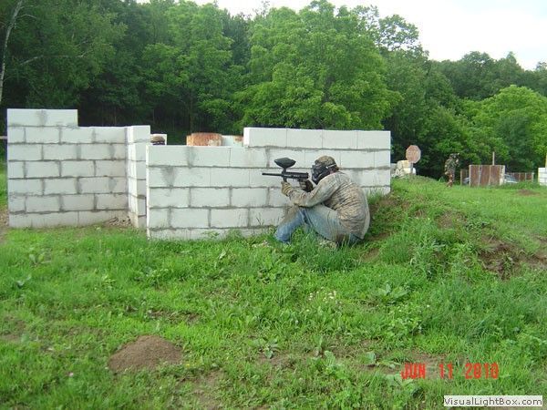 Person crouched with a paintball gun behind a cinder block barricade in a grassy field.