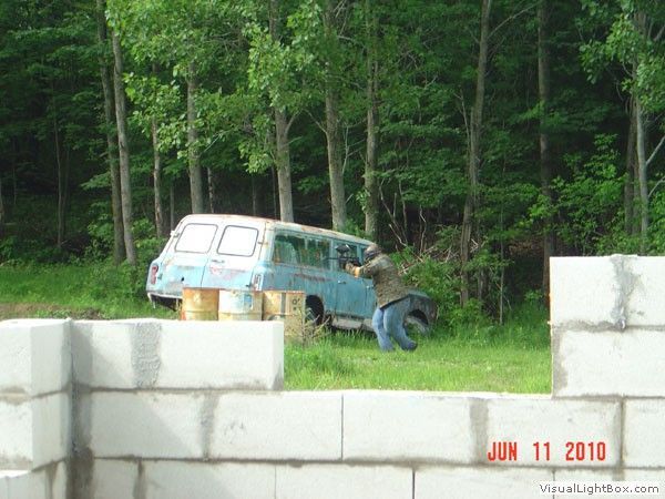 Blue van parked near trees behind a white cinder block wall, with a person bent beside it.