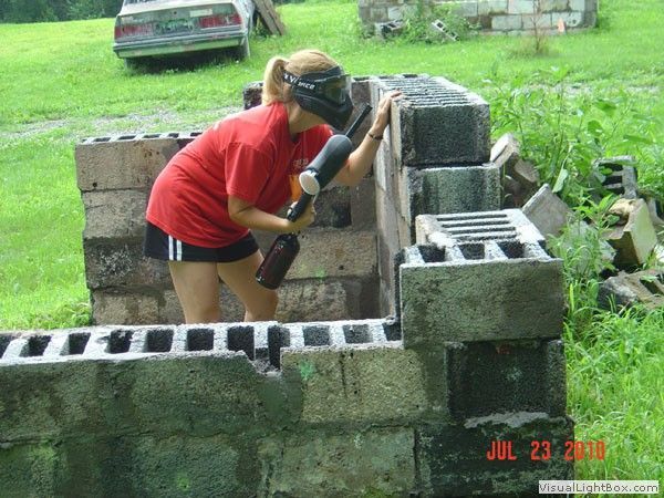 Person in red shirt and black shorts laying cinder blocks outdoors with a hammer in a grassy yard.