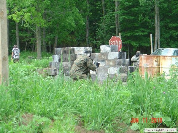 Camouflaged person crouches behind concrete barriers near a stop sign in a wooded area