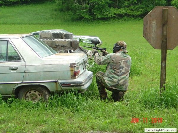 Two people in camouflage crouch behind a white car and aim a rifle in a grassy yard.