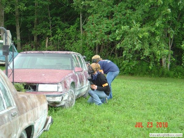 Two people kneel beside a red car in a grassy yard, inspecting the front wheel area.