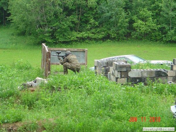 Man in camouflage standing by a small stone memorial in a grassy field near trees