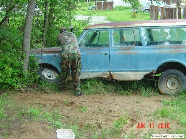Camouflaged person examining a blue station wagon stuck in a muddy yard