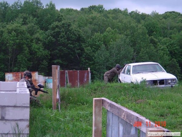 People in camouflage aiming rifles near a white car in a grassy outdoor training area