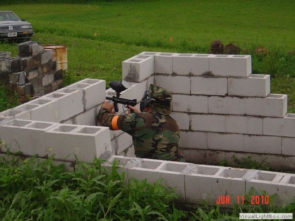 Person in camouflage aiming a rifle from behind a low cinder-block wall in a grassy field.
