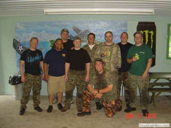 Group of soldiers posing indoors in camouflage uniforms, one kneeling in front of a painted mural backdrop.