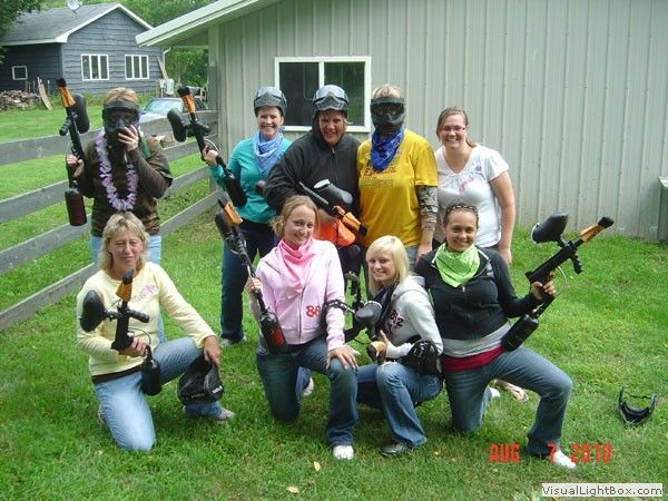 Group posing outdoors with paintball markers, wearing protective masks and colorful gear.