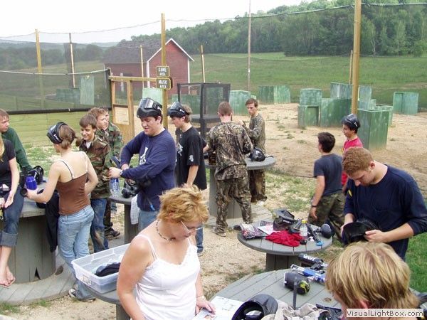 Group of people in camouflage and casual clothes gathered at an outdoor paintball field near barriers and bunkers