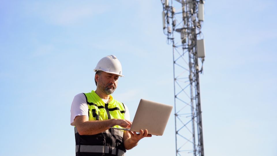 Man in hard hat and vest uses laptop near cell tower.