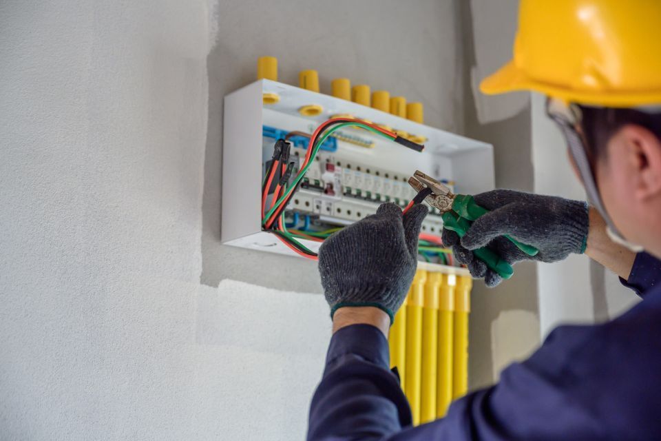 Electrician working on a circuit breaker panel, wearing gloves and a yellow hard hat.