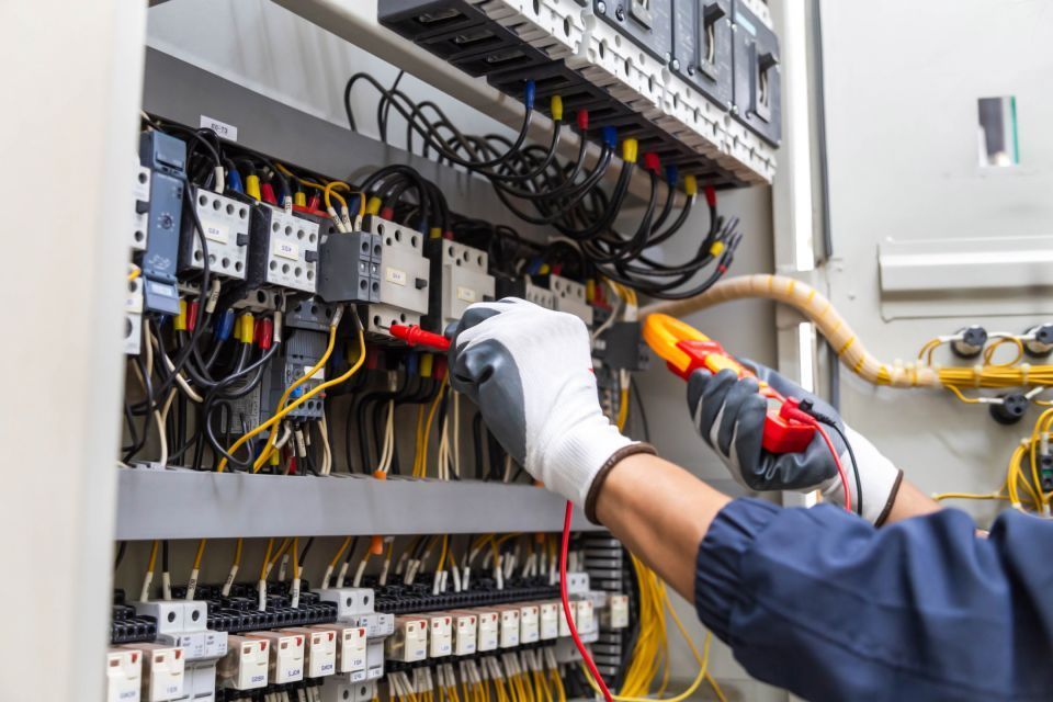 Electrician in gloves testing wires inside a control panel with a multimeter.