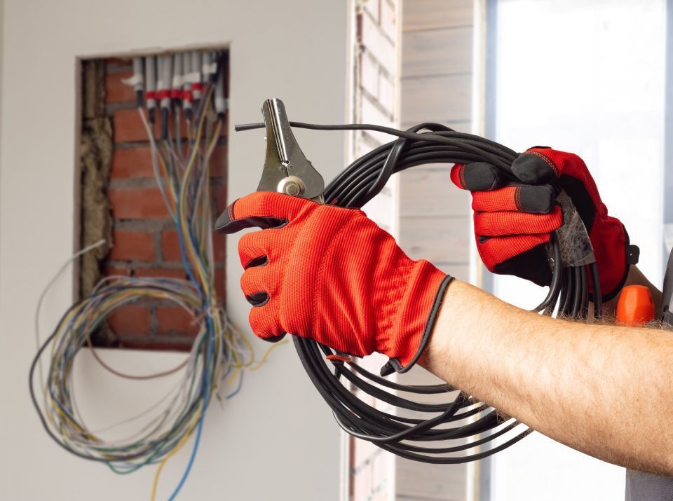 Person wearing red gloves cutting black cable with pliers near exposed electrical wires.