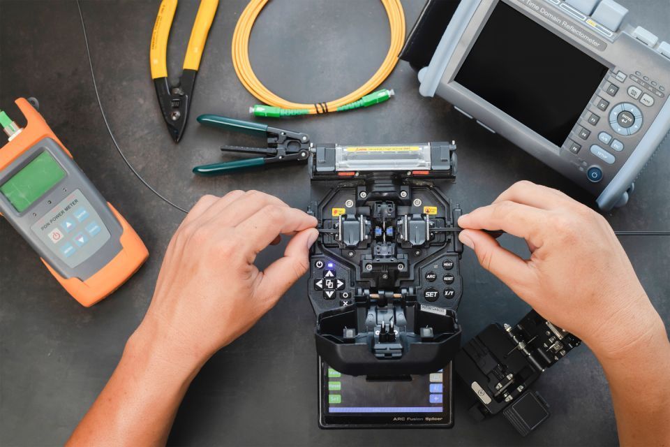 Hands aligning fiber optic cables in a fusion splicer, with tools and testing equipment on a black surface.