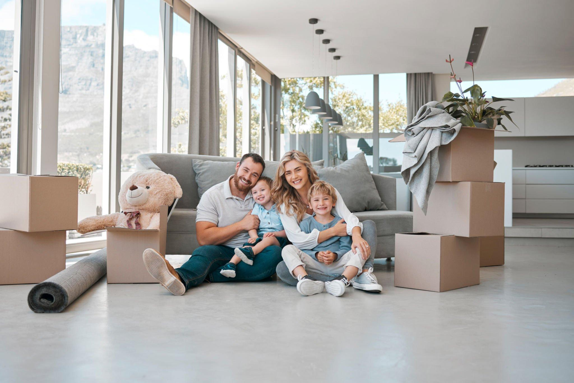 A family is sitting on the floor in a living room surrounded by cardboard boxes.