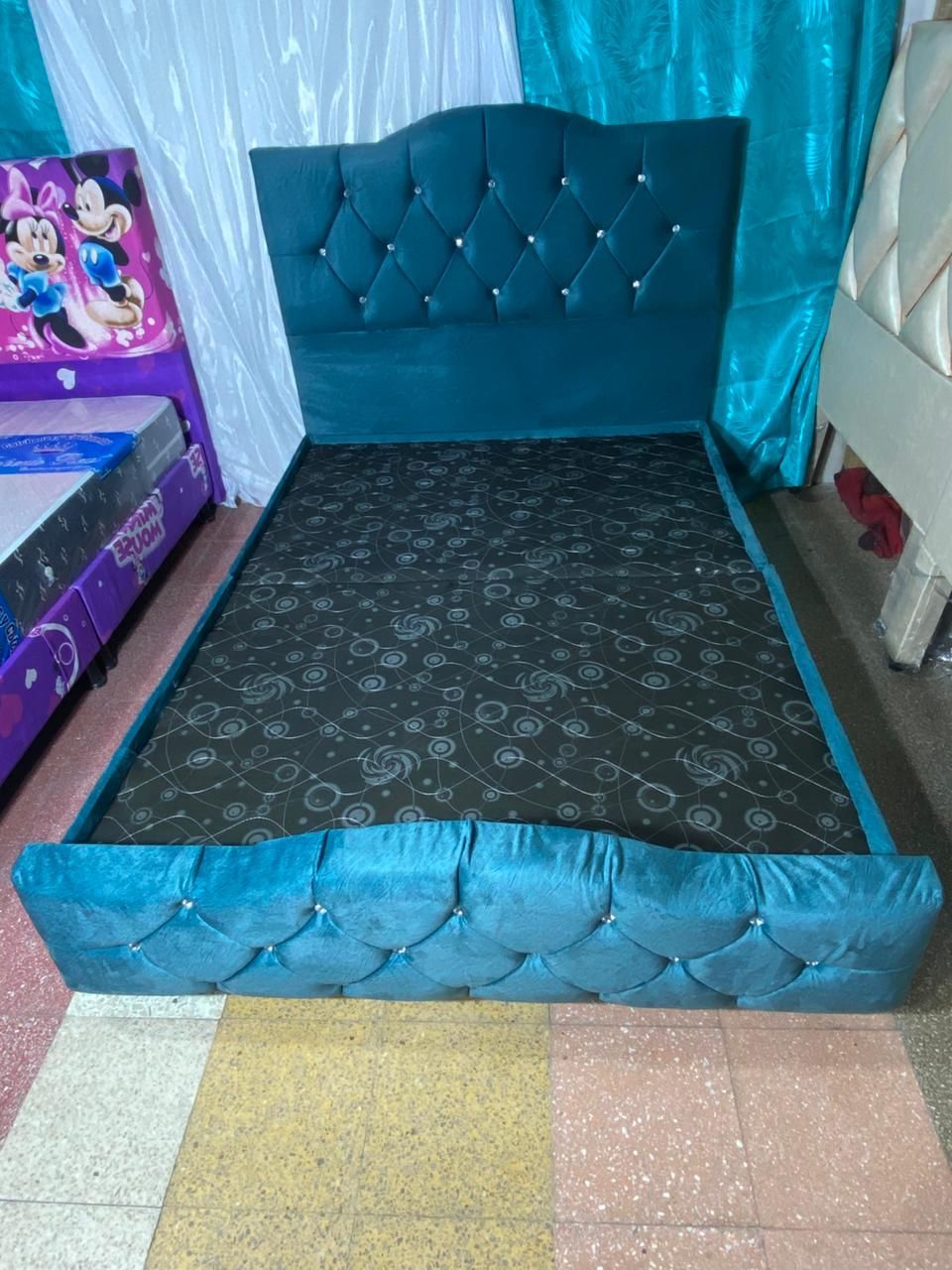 Cama de terciopelo azul con cabecera y pie de cama con mechones capitoné de rombos y colchón con estampado negro.