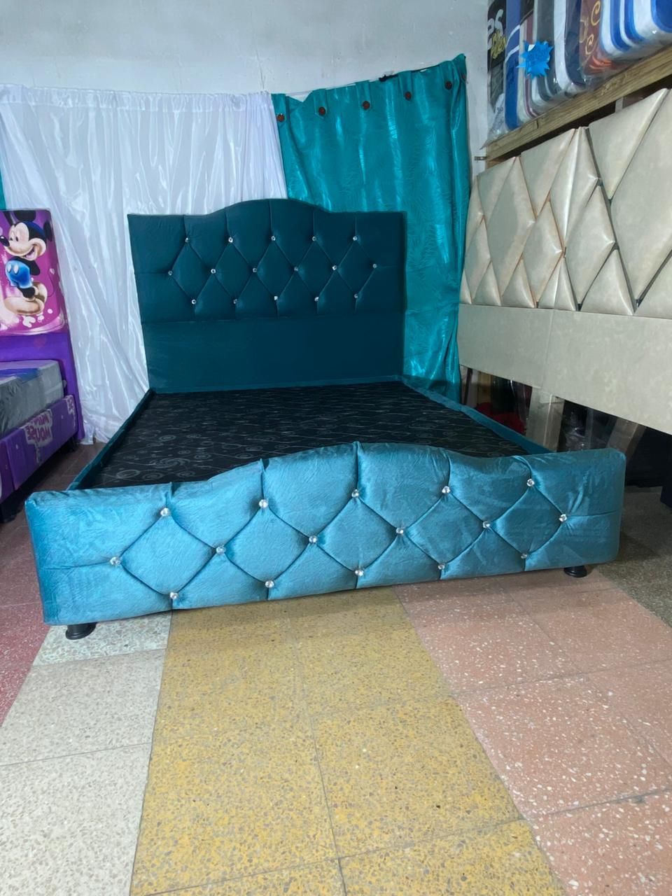 Estructura de cama tapizada en color verde azulado con cabecera y pie de cama capitoné, contrastada con cortinas y otras camas.