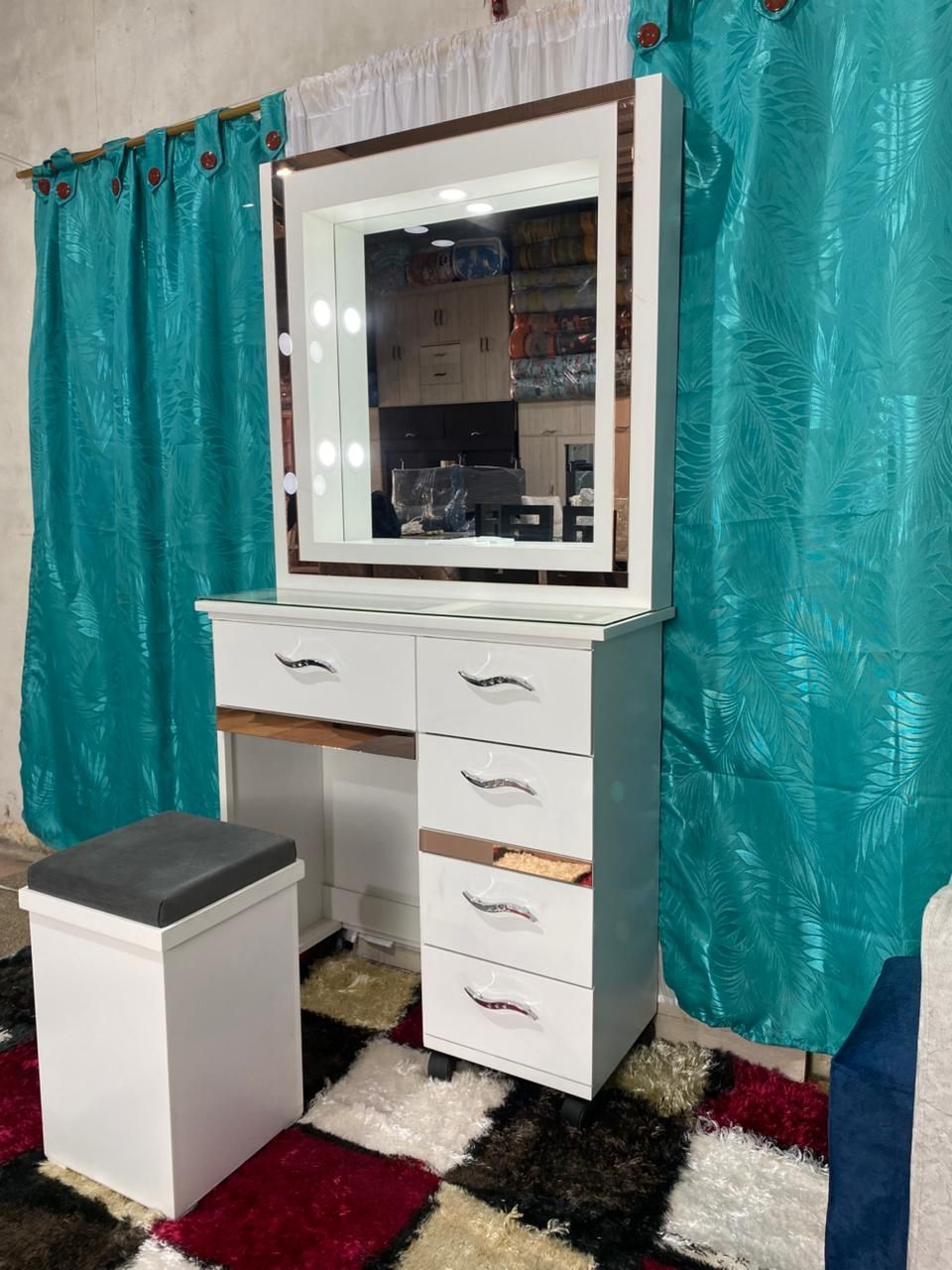 Tocador blanco con espejo, luces y cajones, cortinas turquesas y un taburete sobre una alfombra multicolor.