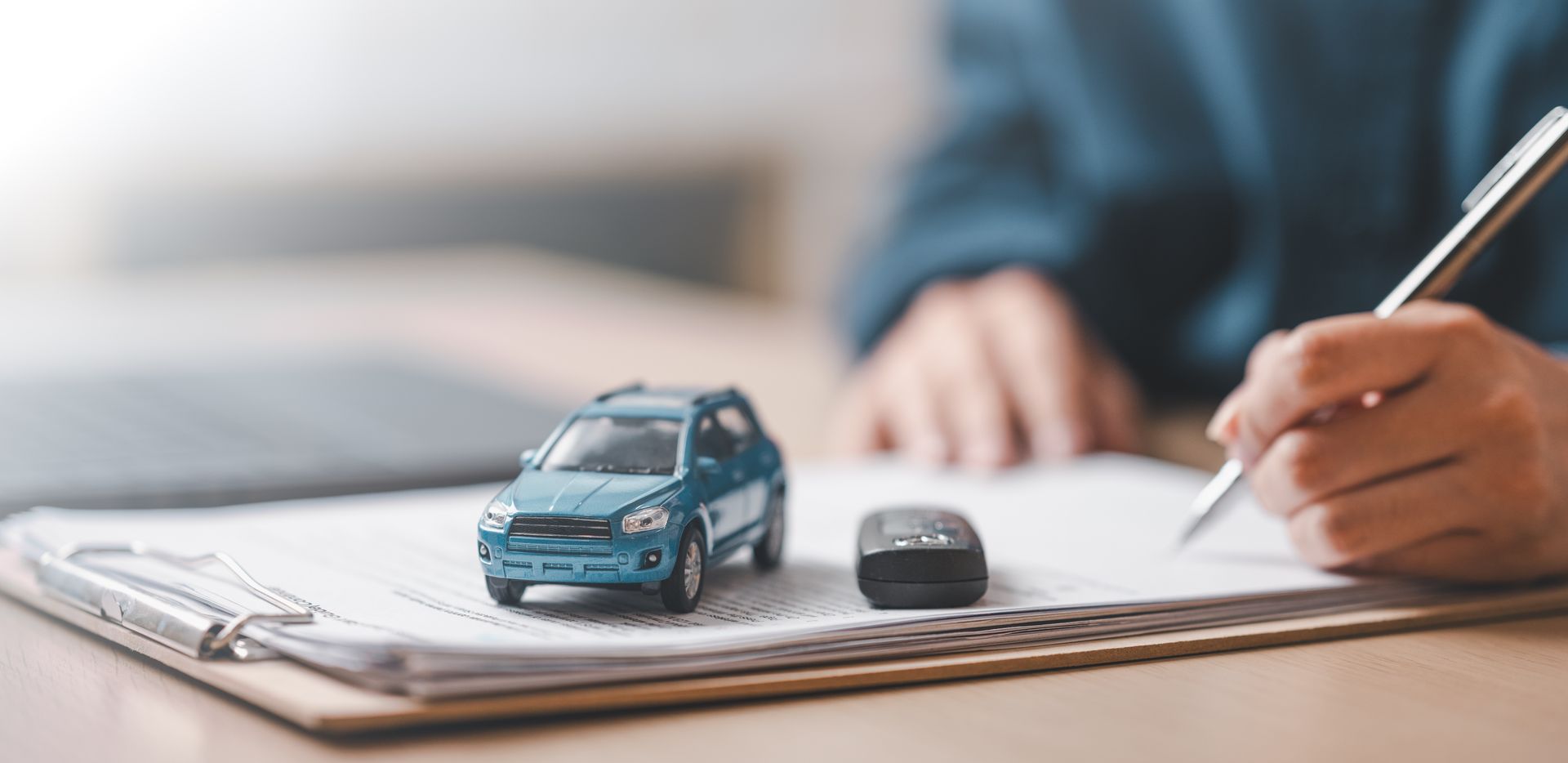 A small model car and key fob rest on documents as a person signs paperwork at a desk.