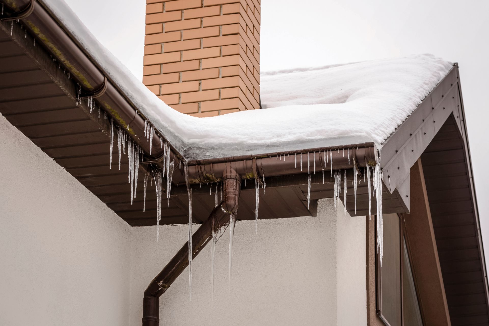 Snow covered roof with icicles hanging from the gutters near a brick chimney.