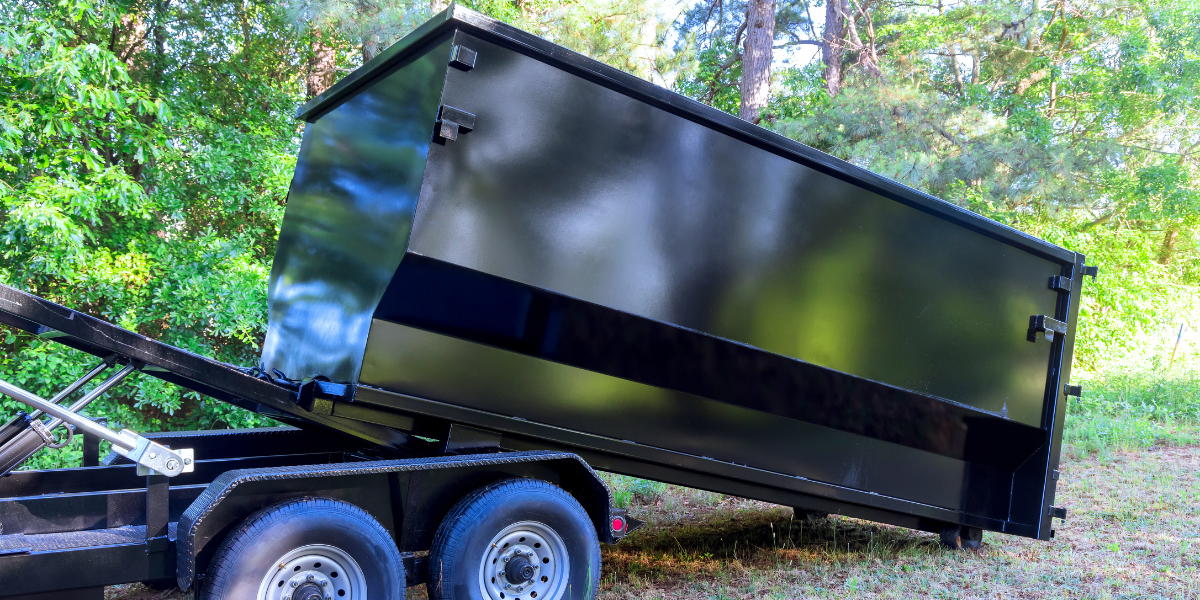 A black dump trailer tipped upward in a wooded area, with the rear of the bin touching the ground.