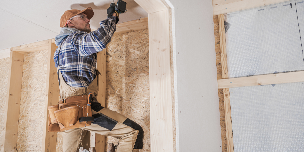 A worker in a plaid shirt and tool belt installs a ceiling fixture in a Big Bear Lake cabin