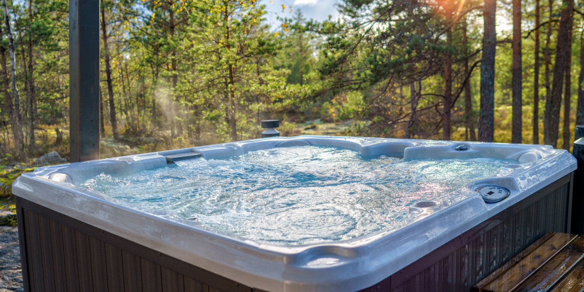 A steaming hot tub sits on a wooden deck surrounded by a sunlit forest in Big Bear