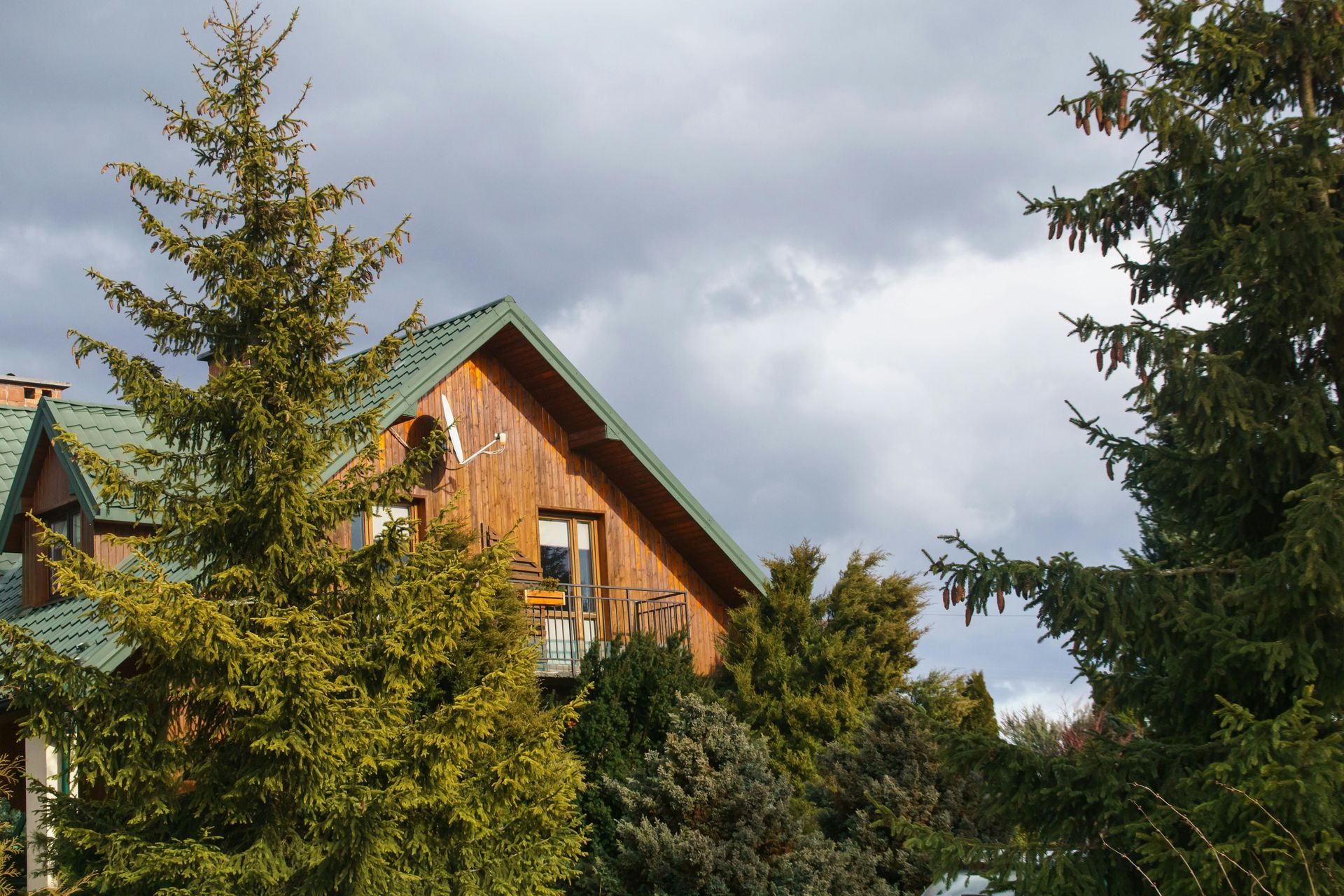 A multi-story wooden house with a green roof nestled among evergreen trees under a cloudy sky.