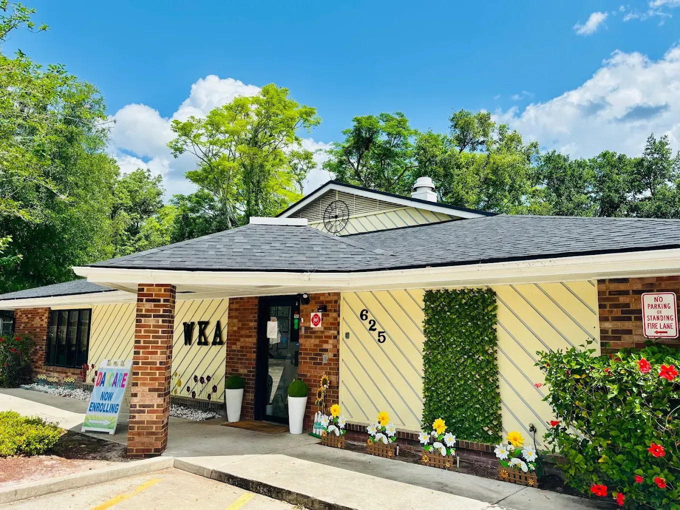 Exterior of a light yellow building with brick columns. Entrance has a sign, door, and potted plants. Lush green trees are in the background.