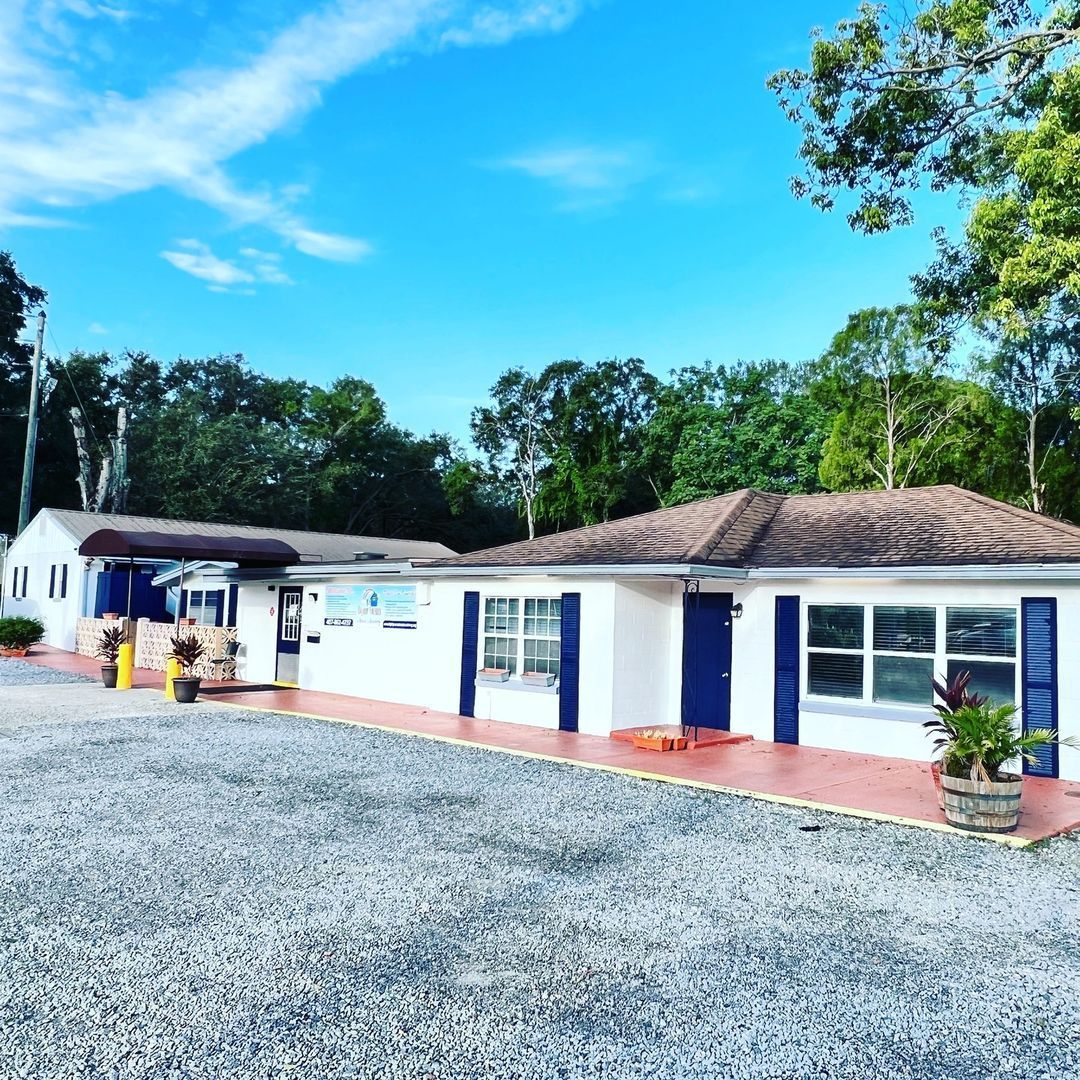 A white house with blue shutters is surrounded by gravel and trees