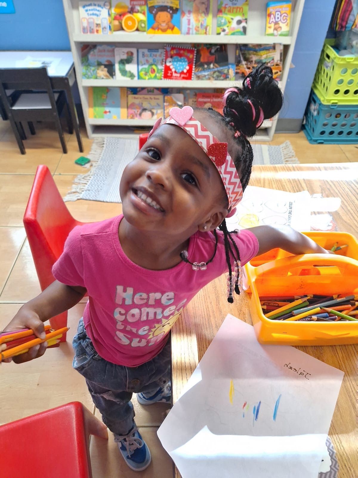 Young girl with a pink shirt, drawing with crayons in a classroom, smiling.