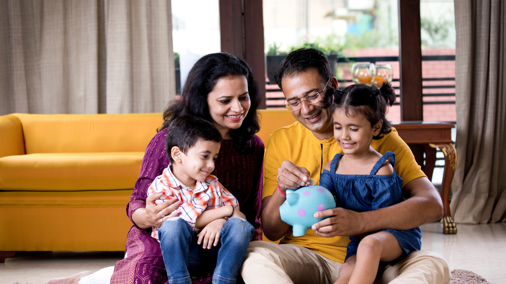 A family is sitting on the floor in a living room putting money into a piggy bank.