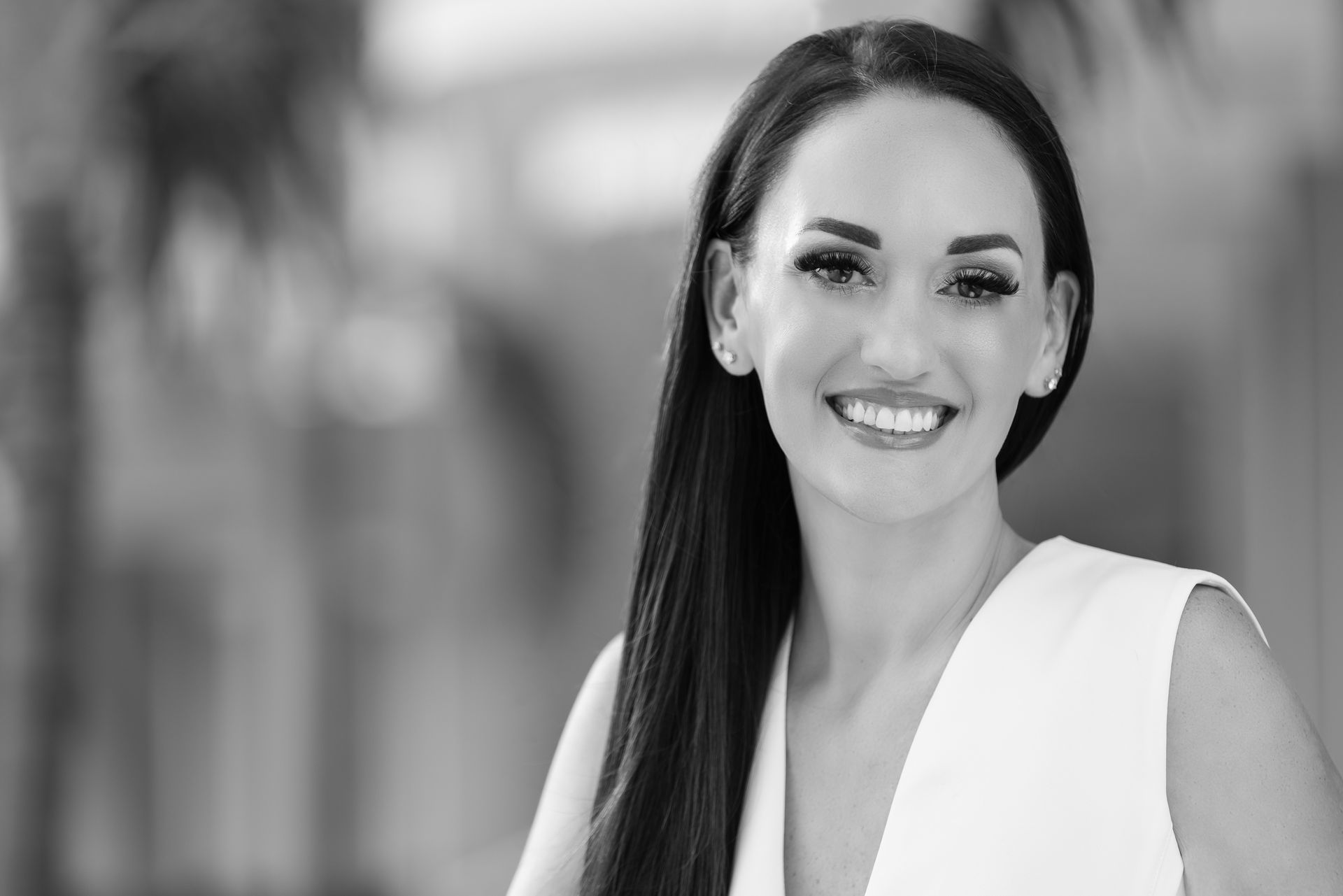 Woman with dark hair smiles, wears white top. Black and white photo, outdoor setting.