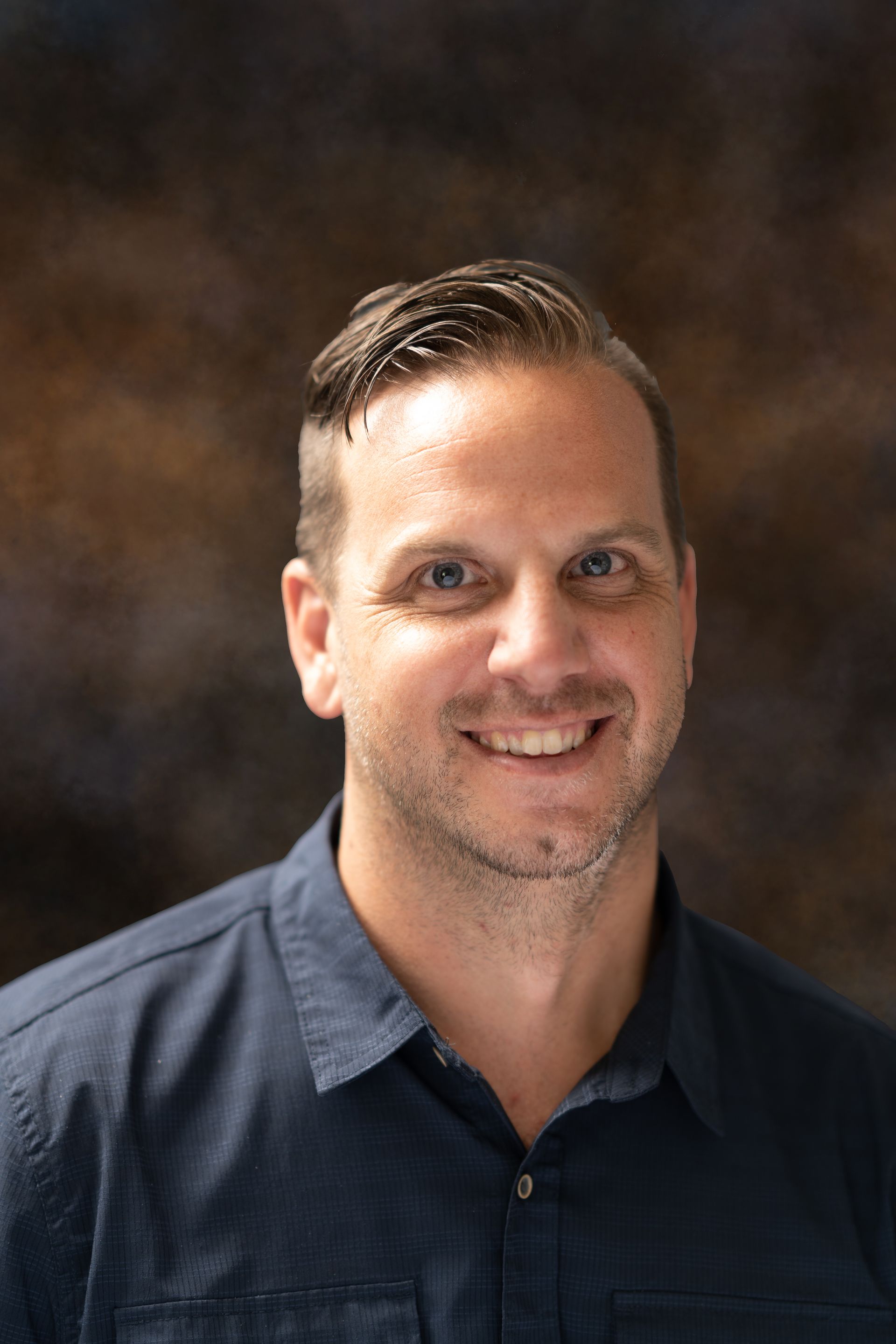 Man with fair skin and short brown hair, smiling, wearing a blue button-down shirt, against a blurry brown background.