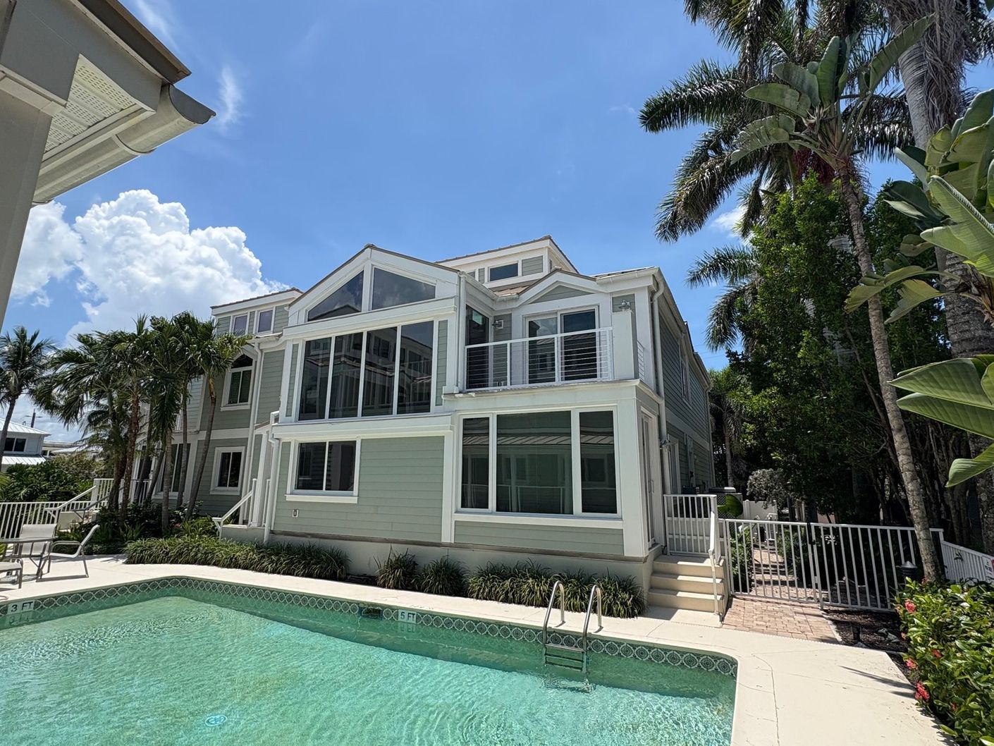 A light green multi-story house with a pool in front; palm trees and a sunny sky.