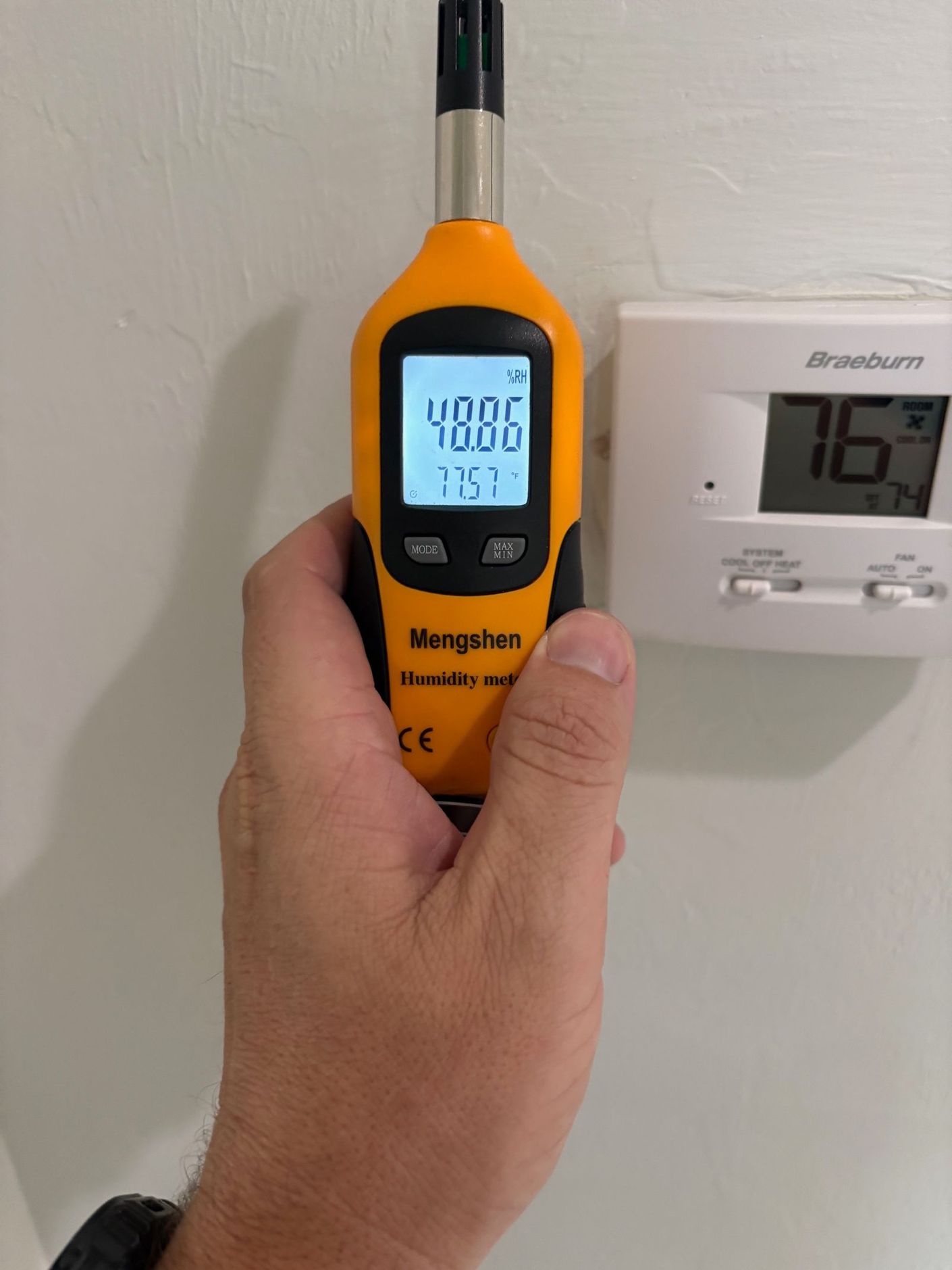 Hand holding a yellow digital hygrometer next to a white thermostat; both are on a white wall.