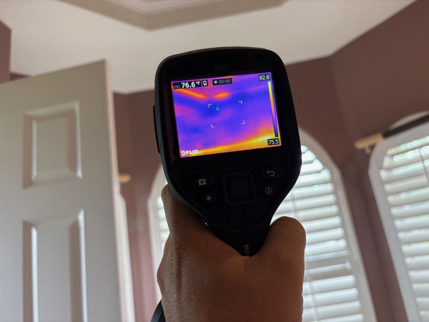 Hand holding a thermal imaging camera, pointing at a ceiling in a room with a door and windows visible.