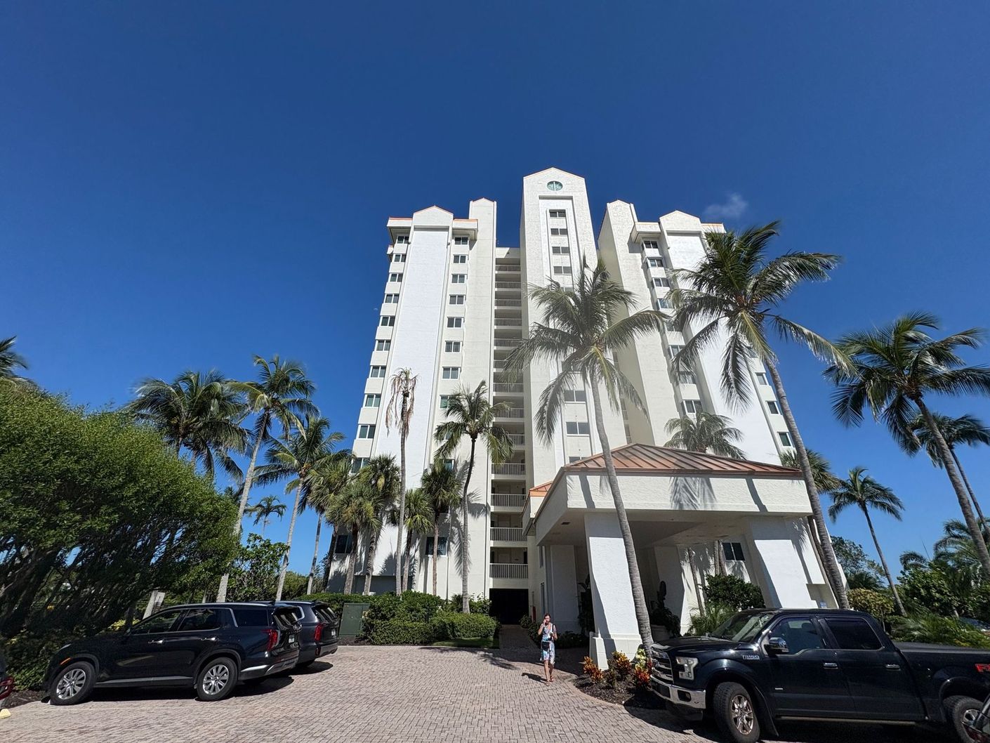 White high-rise building with palm trees, cars parked in front, against a blue sky.