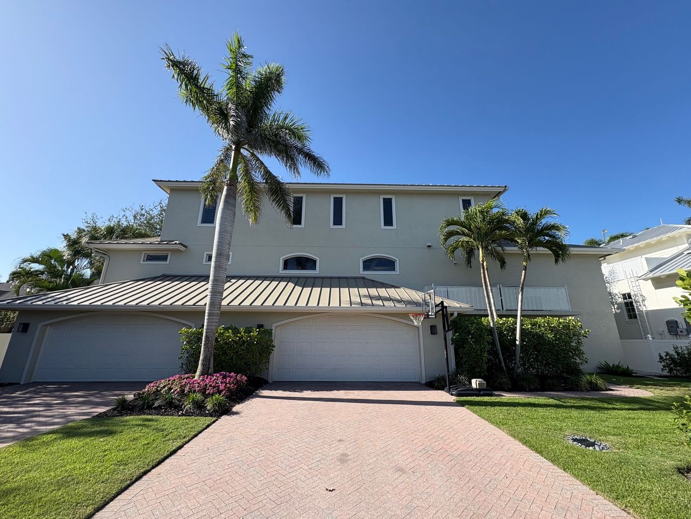 Two-story gray house with garage, driveway, palm trees, and blue sky.