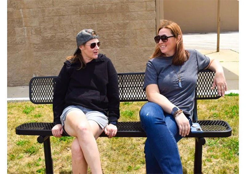 Two women sitting on a bench, smiling and talking. One wears a cap and black hoodie, the other sunglasses and a gray shirt.