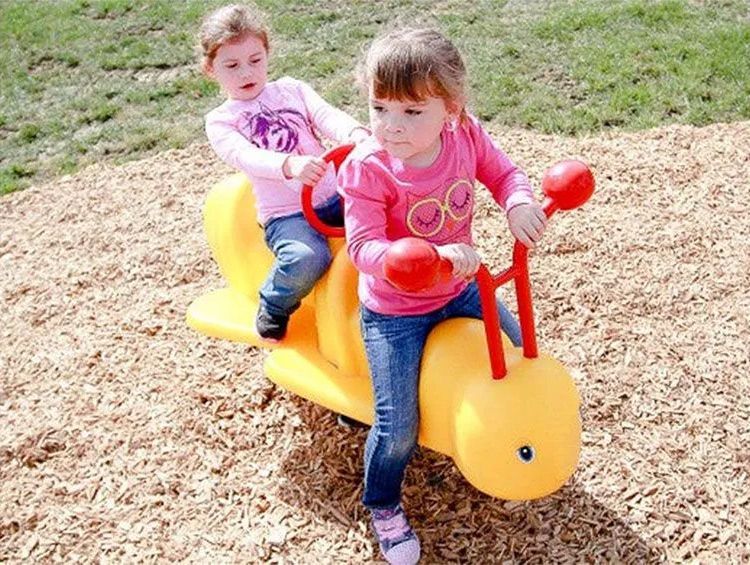 Two children play on a yellow bug-shaped playground teeter-totter on wood chips.
