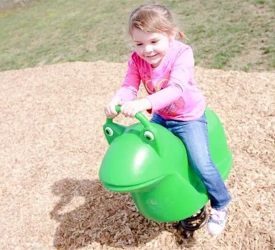 Smiling child rides a green spring-mounted frog toy at a playground on wood chips.
