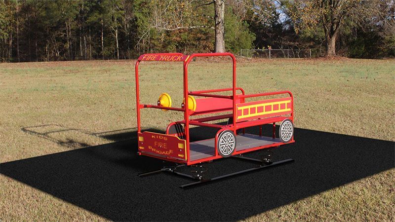 Fire truck-themed playground spinner on black mat in grassy field. Red, yellow, and silver.