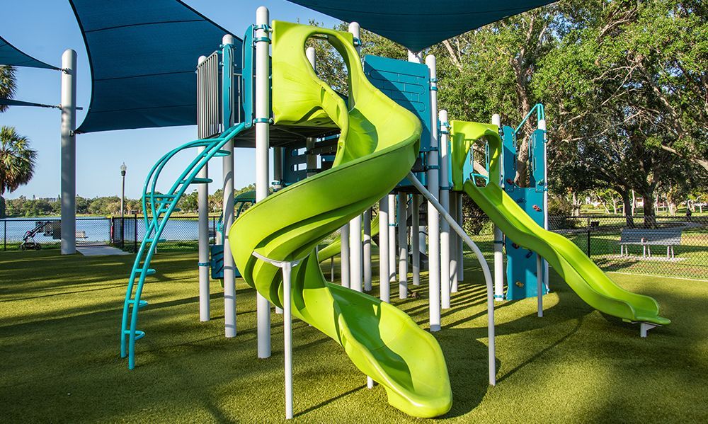 Playground with green slides, teal ladder, and blue shade in park setting.