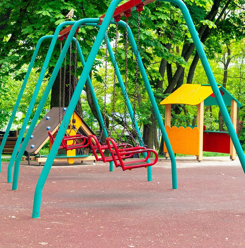 Swingset in a playground with colorful structures and trees in the background.