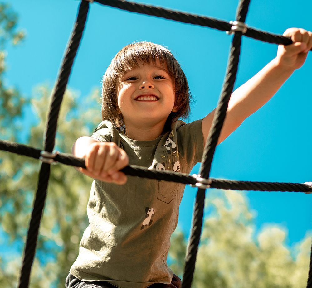Child smiling, climbing a black rope net against a bright blue sky.