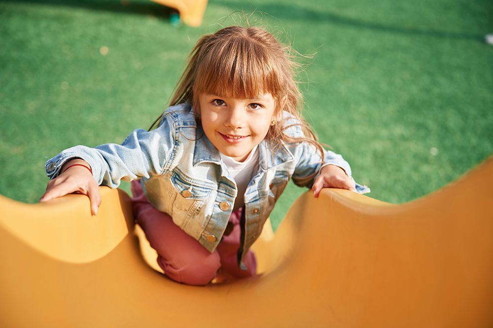Girl smiles while climbing up a yellow slide on a green play area.