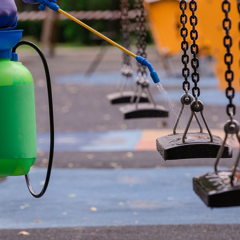 Person sanitizing playground swings with a sprayer. Green tank and blue nozzle.