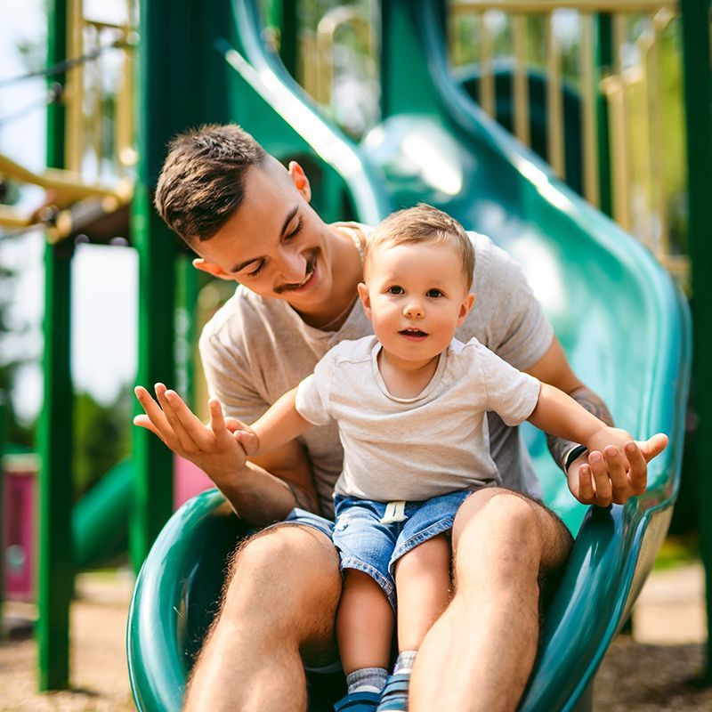 Man and toddler on a green slide at a playground; man smiles, toddler looks forward.