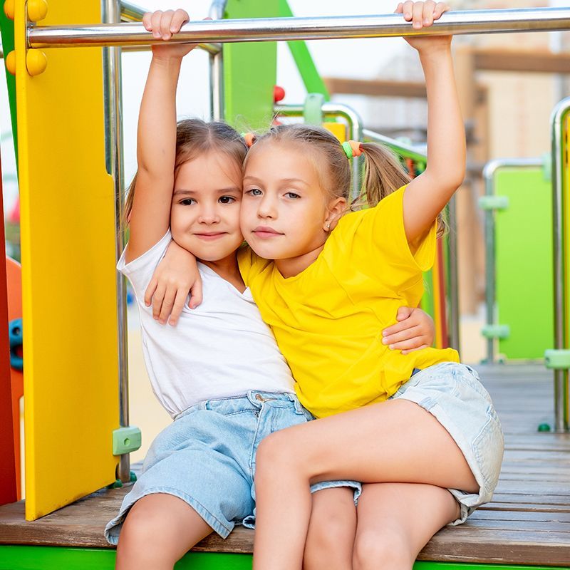 Two girls hugging, smiling, sitting on playground structure. One wears yellow, the other white. Bright colors.